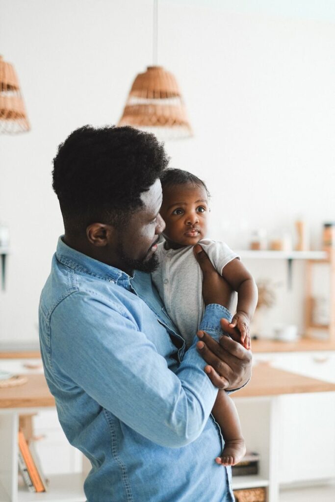 An African American father lovingly holds his baby in a cozy, well-lit kitchen setting.