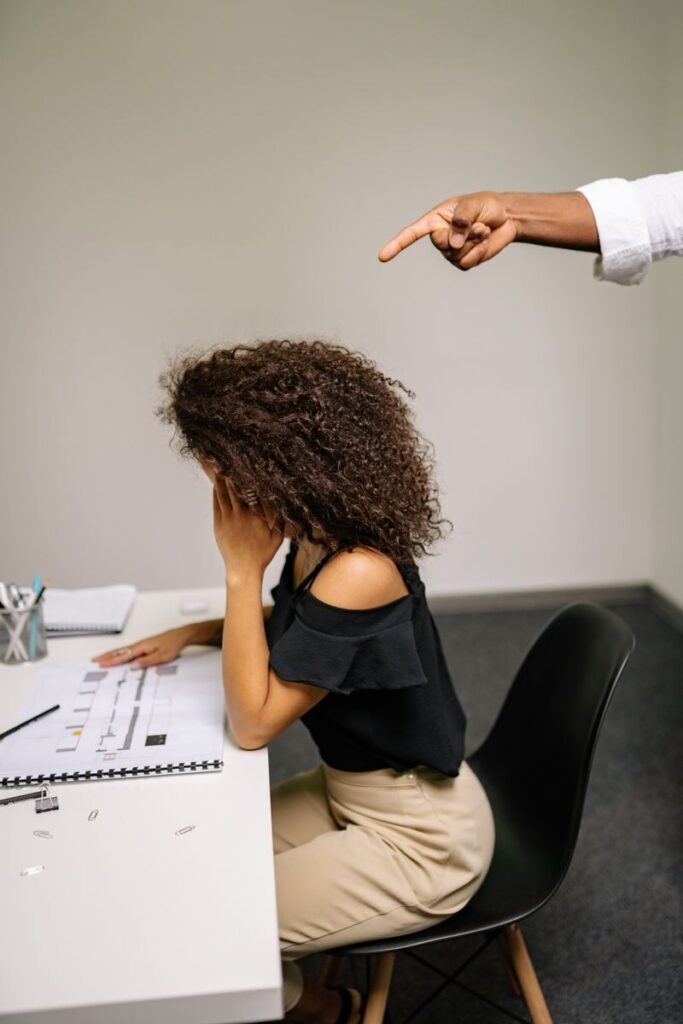 A stressed woman sits at her desk while a hand points at her, depicting office tension.
